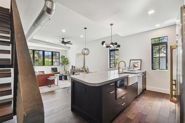 kitchen with an island with sink, dark hardwood / wood-style flooring, stainless steel dishwasher, ceiling fan with notable chandelier, and decorative light fixtures