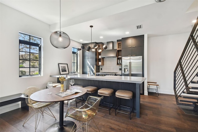 kitchen with tasteful backsplash, decorative light fixtures, dark wood-type flooring, stainless steel fridge with ice dispenser, and a center island with sink