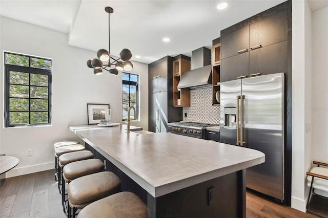 kitchen featuring wall chimney range hood, dark hardwood / wood-style flooring, a breakfast bar area, a kitchen island with sink, and high end appliances