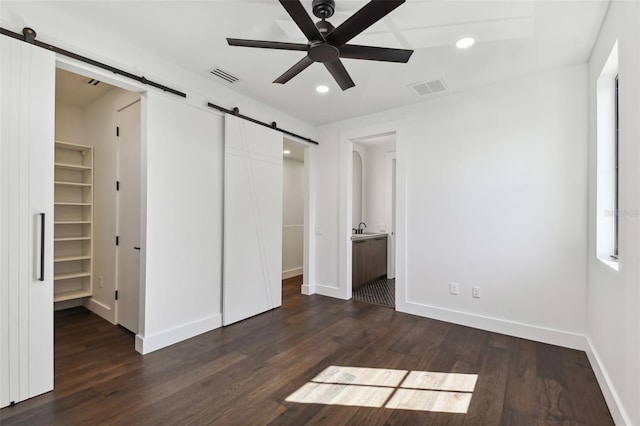 unfurnished bedroom featuring dark wood-type flooring, a spacious closet, a barn door, ensuite bathroom, and ceiling fan