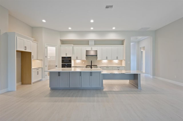 kitchen with light hardwood / wood-style flooring, a kitchen island with sink, backsplash, and white cabinetry