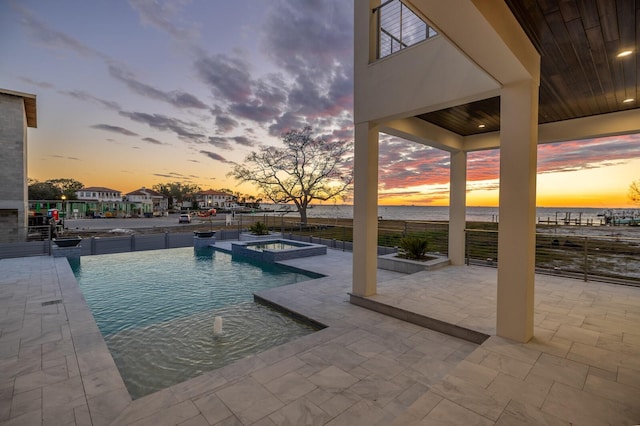 pool at dusk with an in ground hot tub, a water view, and a patio area
