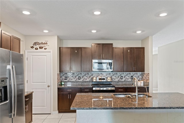 kitchen with dark stone counters, stainless steel appliances, sink, light tile patterned floors, and a center island with sink