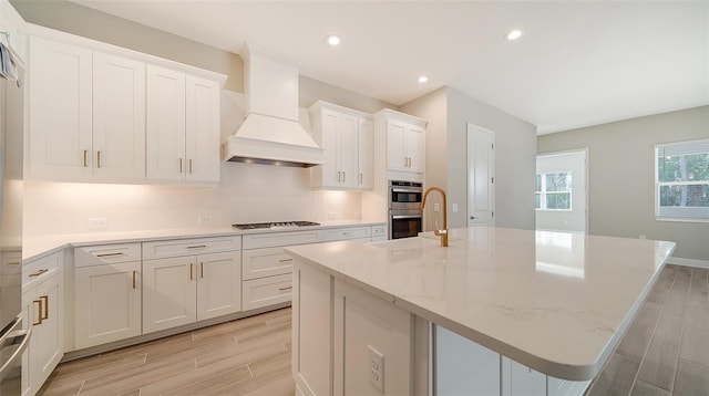 kitchen featuring gas cooktop, premium range hood, double oven, an island with sink, and white cabinets