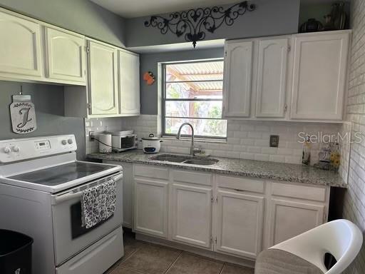 kitchen with white range with electric stovetop, tile patterned floors, a sink, white cabinetry, and backsplash