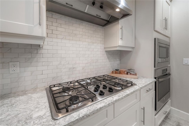 kitchen featuring wall chimney exhaust hood, white cabinetry, light stone counters, tasteful backsplash, and stainless steel appliances