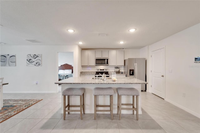 kitchen featuring appliances with stainless steel finishes, a center island with sink, light stone counters, light tile patterned floors, and a kitchen breakfast bar