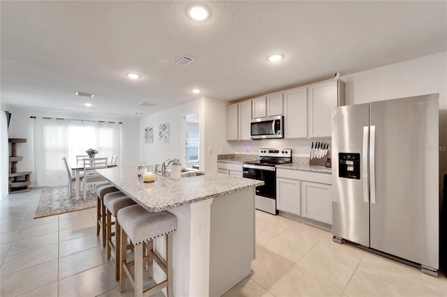 kitchen featuring light tile patterned flooring, stainless steel appliances, sink, light stone countertops, and a center island with sink