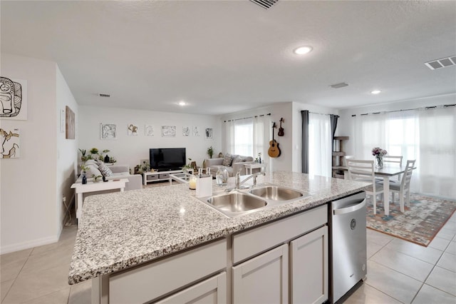 kitchen with stainless steel dishwasher, light tile patterned flooring, sink, light stone counters, and a kitchen island with sink