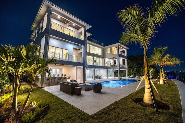 back house at night featuring an outdoor living space, a balcony, ceiling fan, a lawn, and a patio area