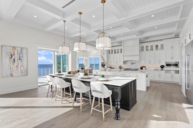 kitchen with light wood-type flooring, a large island with sink, beam ceiling, hanging light fixtures, and a water view