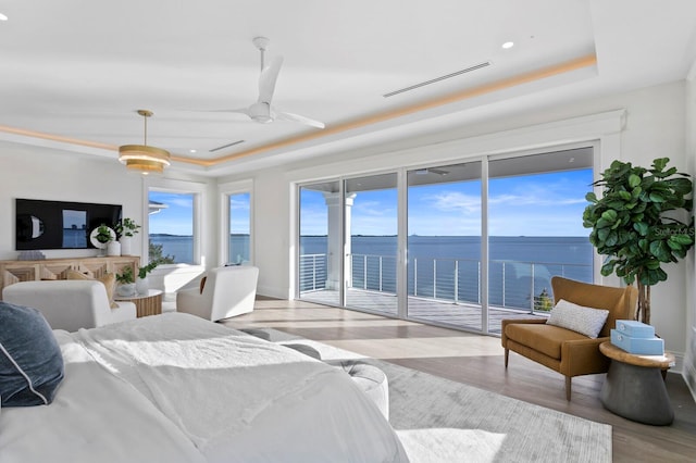 bedroom featuring a water view, access to exterior, ceiling fan, a raised ceiling, and light wood-type flooring