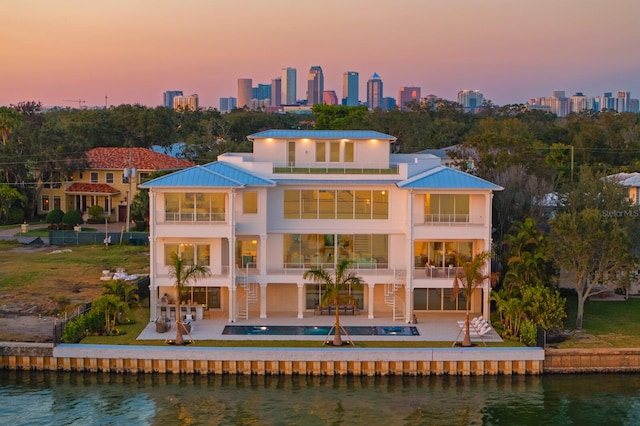 back house at dusk with a balcony, a water view, a swimming pool with hot tub, and a patio area