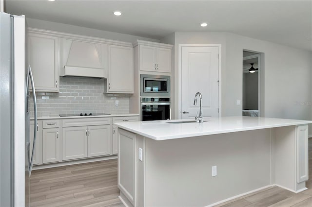 kitchen featuring a kitchen island with sink, premium range hood, light wood-type flooring, and stainless steel appliances