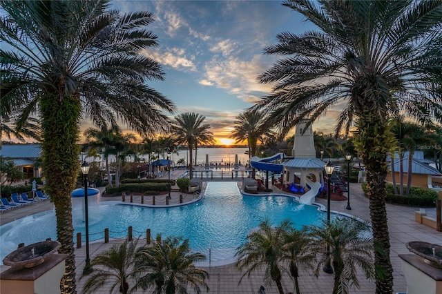 pool at dusk featuring a water view, a water slide, and a patio