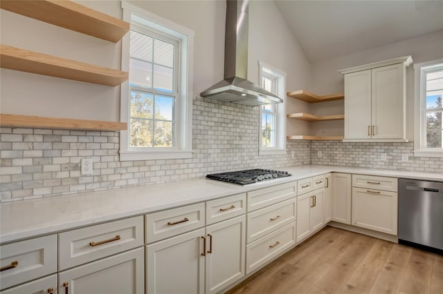 kitchen featuring appliances with stainless steel finishes, light hardwood / wood-style flooring, vaulted ceiling, wall chimney exhaust hood, and tasteful backsplash