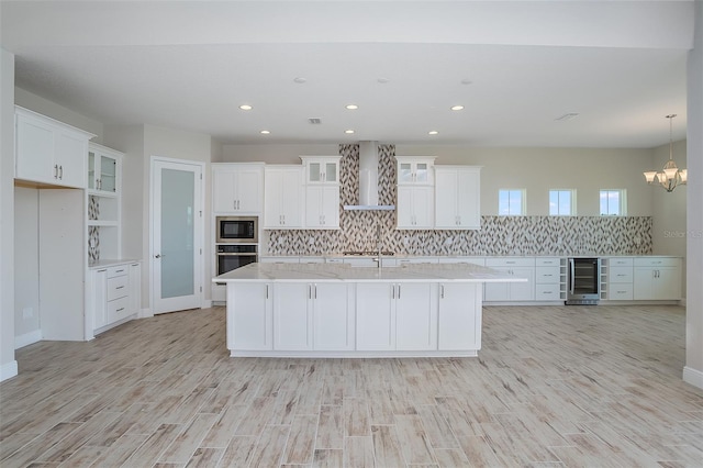 kitchen featuring beverage cooler, stainless steel appliances, wall chimney range hood, a center island with sink, and white cabinets