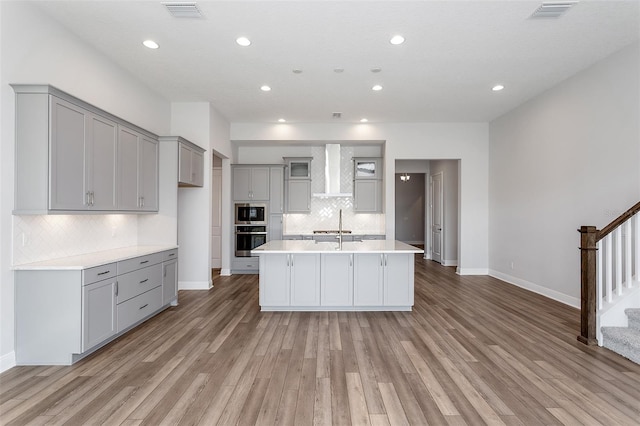 kitchen with gray cabinetry, stainless steel microwave, backsplash, light hardwood / wood-style flooring, and an island with sink