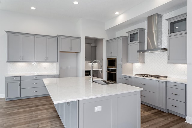 kitchen with stainless steel appliances, a kitchen island with sink, dark wood-type flooring, sink, and wall chimney range hood