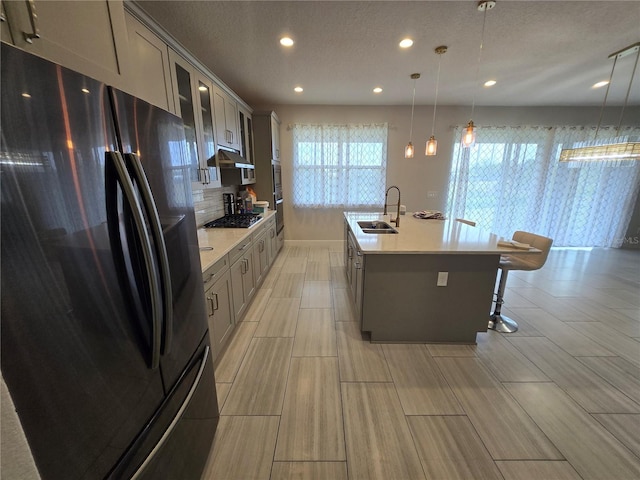 kitchen featuring stainless steel gas cooktop, a center island with sink, light countertops, freestanding refrigerator, and a sink