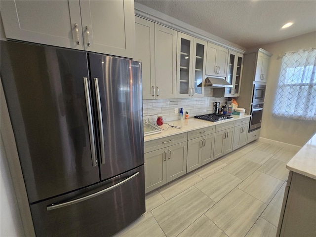 kitchen featuring tasteful backsplash, glass insert cabinets, under cabinet range hood, gray cabinets, and appliances with stainless steel finishes