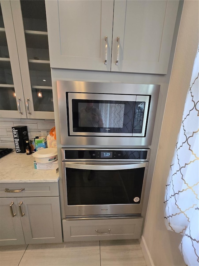 room details featuring tasteful backsplash, glass insert cabinets, light stone countertops, gray cabinets, and stainless steel appliances