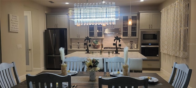 kitchen with an island with sink, under cabinet range hood, glass insert cabinets, appliances with stainless steel finishes, and decorative backsplash