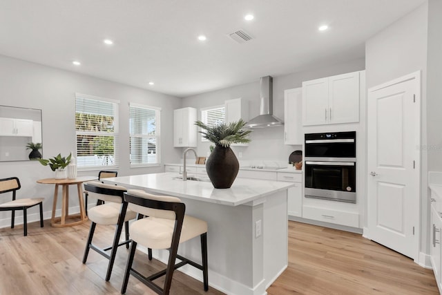 kitchen with wall chimney range hood, sink, white cabinetry, black appliances, and a center island with sink