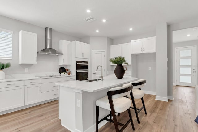 kitchen with white cabinetry, sink, a kitchen island with sink, and wall chimney range hood