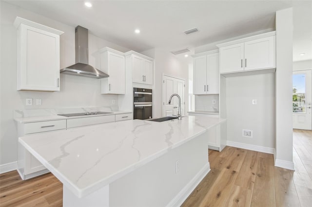 kitchen with wall chimney exhaust hood, black appliances, a kitchen island with sink, and white cabinetry