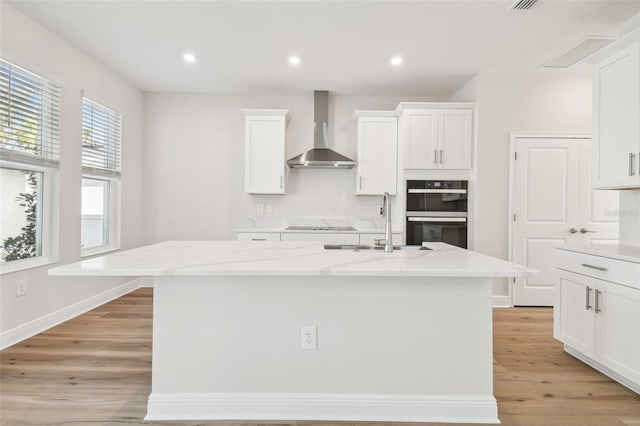 kitchen with wall chimney exhaust hood, white cabinets, light stone countertops, black double oven, and a center island with sink