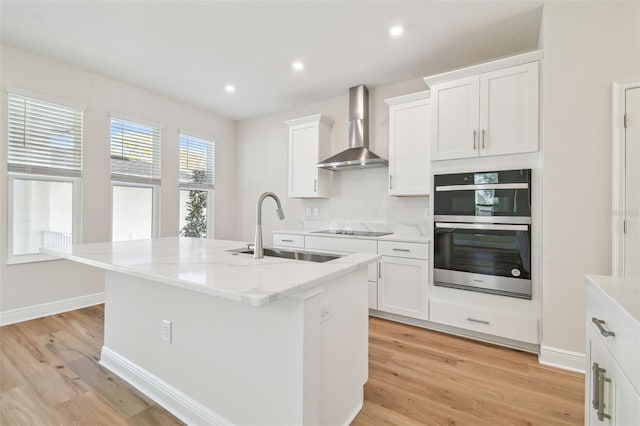 kitchen with sink, white cabinetry, a kitchen island with sink, wall chimney range hood, and double wall oven