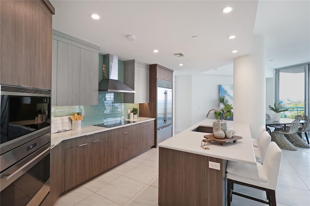 kitchen with wall chimney range hood, built in appliances, an island with sink, a kitchen bar, and sink