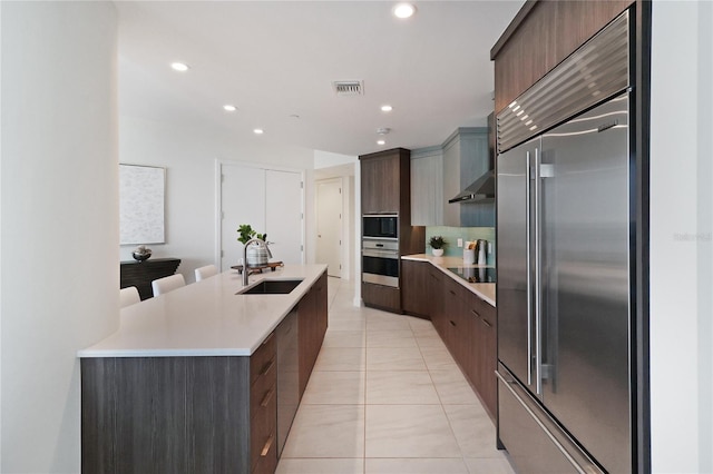 kitchen with dark brown cabinetry, a kitchen island with sink, built in refrigerator, sink, and light tile patterned flooring