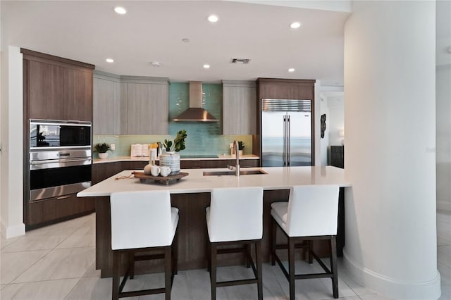 kitchen with appliances with stainless steel finishes, a kitchen island with sink, wall chimney exhaust hood, and backsplash