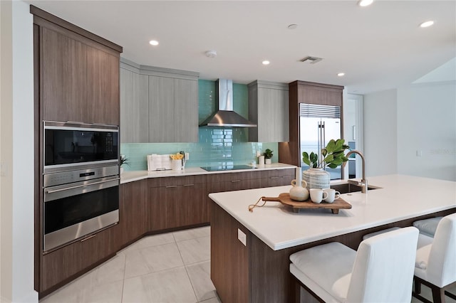 kitchen with appliances with stainless steel finishes, decorative backsplash, wall chimney range hood, and an island with sink