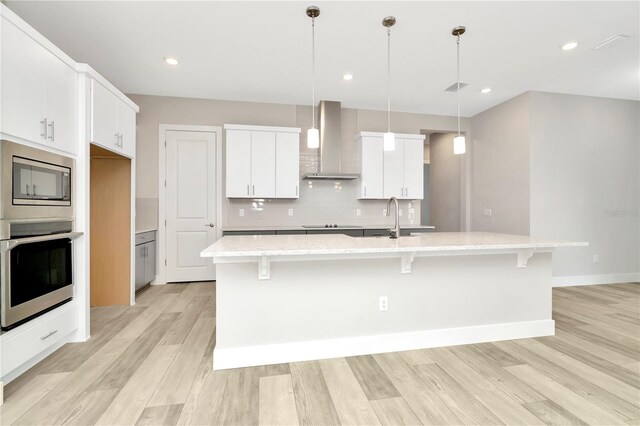 kitchen featuring appliances with stainless steel finishes, white cabinetry, a kitchen island with sink, and wall chimney range hood