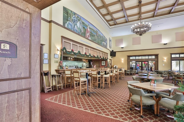 carpeted dining area featuring indoor bar, coffered ceiling, a high ceiling, beam ceiling, and an inviting chandelier