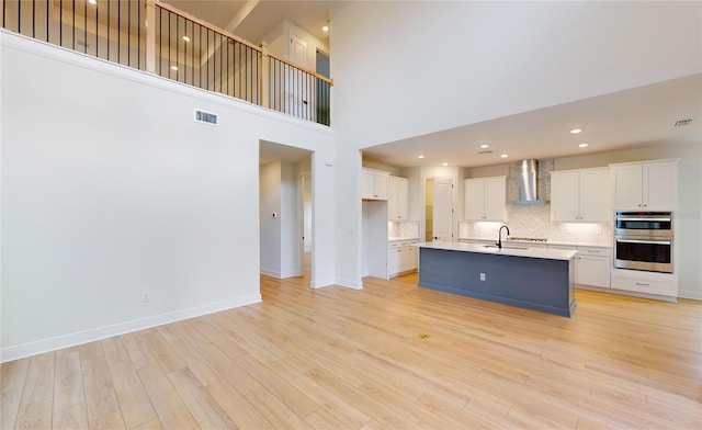 kitchen featuring wall chimney exhaust hood, stainless steel double oven, a kitchen island with sink, white cabinets, and a high ceiling