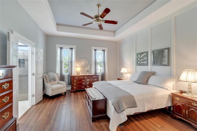 bedroom with a tray ceiling, dark wood-type flooring, ceiling fan, and ensuite bath