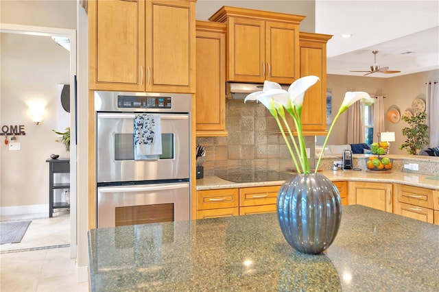 kitchen featuring ceiling fan, light tile floors, dark stone counters, double oven, and tasteful backsplash