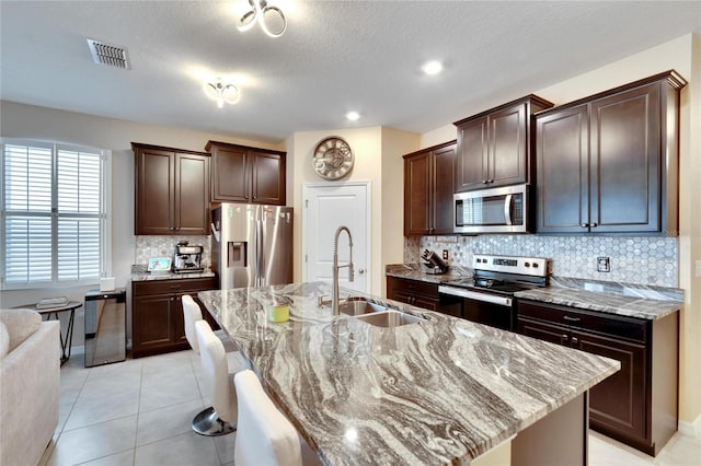 kitchen featuring a center island with sink, stainless steel appliances, visible vents, a sink, and dark brown cabinetry