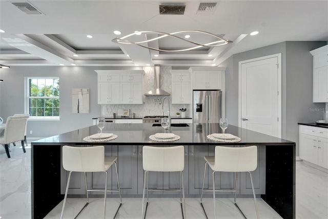 kitchen with stainless steel fridge with ice dispenser, light tile flooring, wall chimney exhaust hood, an island with sink, and a raised ceiling