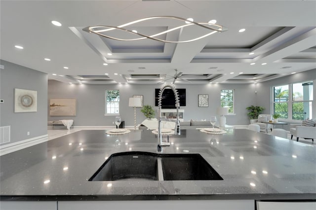 kitchen with coffered ceiling, sink, dark stone counters, an island with sink, and an inviting chandelier