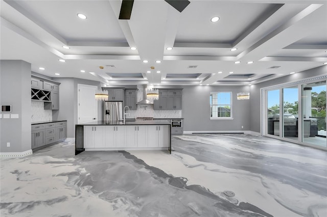 kitchen with coffered ceiling, an island with sink, tasteful backsplash, wall chimney exhaust hood, and gray cabinetry