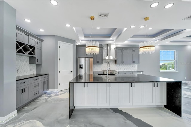 kitchen featuring a tray ceiling, hanging light fixtures, an island with sink, and wall chimney range hood