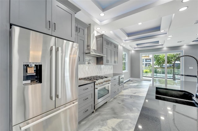 kitchen featuring dark stone counters, a raised ceiling, stainless steel appliances, wall chimney exhaust hood, and tasteful backsplash