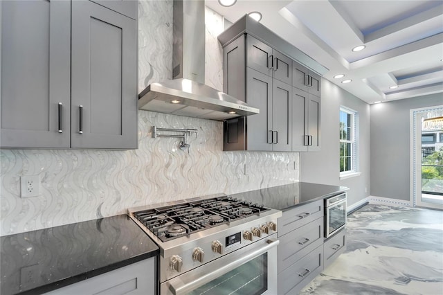 kitchen with dark stone counters, a raised ceiling, wall chimney exhaust hood, tasteful backsplash, and high end stainless steel range oven