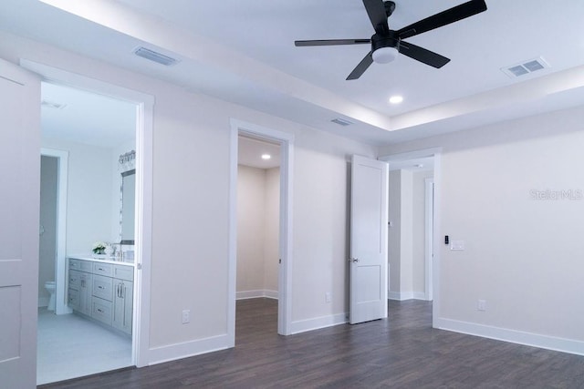 unfurnished bedroom featuring ensuite bathroom, ceiling fan, dark wood-type flooring, and a tray ceiling