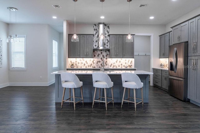 kitchen featuring a center island with sink, decorative light fixtures, stainless steel fridge with ice dispenser, and wall chimney range hood